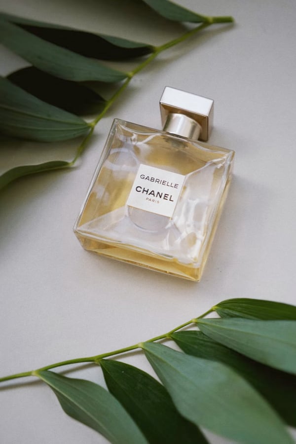 Bottle of Gabrielle Essence Eau De Parfum by Chanel Paris laid out on a cream surface with dark green leaves.