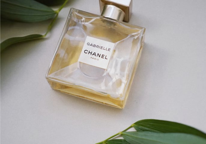 Bottle of Gabrielle Essence Eau De Parfum by Chanel Paris laid out on a cream surface with dark green leaves.